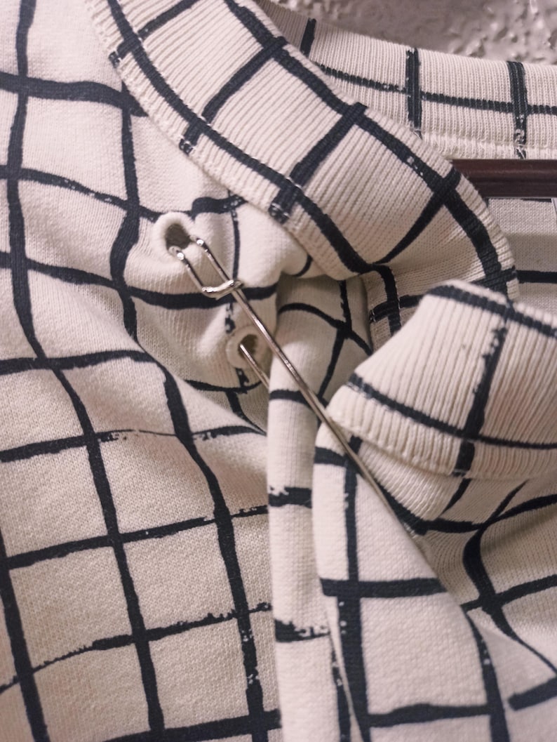 May include: Close-up of a cream-colored sweater with a black grid pattern. A silver safety pin is attached to the collar. The sweater is hanging on a wooden hanger.