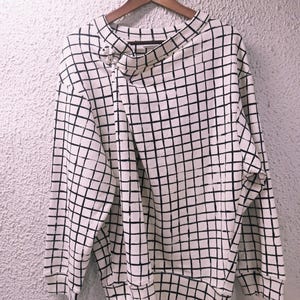 May include: A white sweatshirt with a black grid pattern. The long-sleeved top has a unique asymmetrical neckline and is hanging on a wooden hanger. The fabric appears to be a medium weight, suitable for cooler weather.