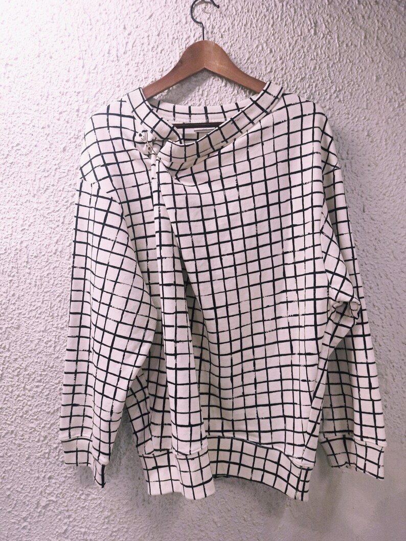 May include: A white sweatshirt with a black grid pattern. The long-sleeved top has a unique asymmetrical neckline and is hanging on a wooden hanger. The fabric appears to be a medium weight, suitable for cooler weather.