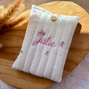Peut inclure: Pochette en tissu blanc rayé, brodée du prénom "Julie" en rose avec des motifs floraux. Un bouton en bois ferme le rabat. La pochette est posée sur un plateau en bois.