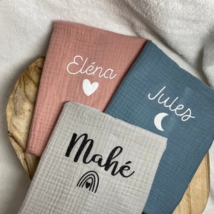 May include: Three personalised baby blankets in pink, blue, and grey. Each blanket features a name and a small graphic. The pink blanket has the name "Éléna" with a heart, the blue blanket has "Jules" with a moon, and the grey blanket has "Mahé" with a rainbow.