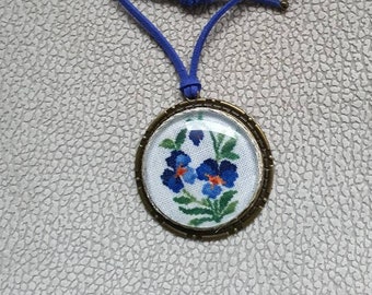 Médaillon en bronze fleurs bleues, pensées en broderie fine sous cabochon. Pendentif fleur. Lacet en cuir réglable.