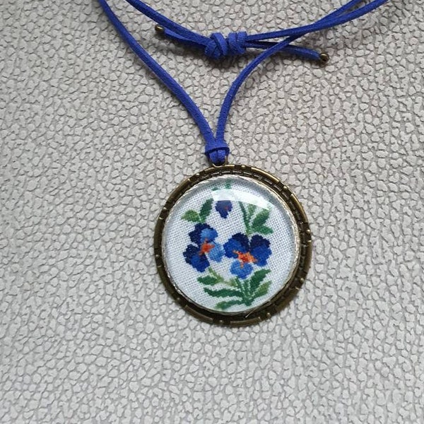 Médaillon en bronze fleurs bleues, pensées en broderie fine sous cabochon. Pendentif fleur. Lacet en cuir réglable.