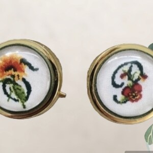 Può includere: Due scatole rotonde decorative con bordo dorato e interno in tessuto bianco. La scatola a sinistra presenta un disegno a punto croce di un girasole. La scatola a destra presenta un disegno a punto croce di una lettera 'S' con un fiore.