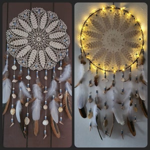 Puede incluir: Un atrapasueños de crochet blanco con plumas blancas y cuentas azules. El atrapasueños está colgado de una pared de madera y tiene una luz que brilla a través de él.