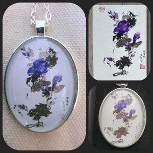 May include: A silver pendant necklace with a round glass charm featuring a watercolor painting of purple flowers on a white background. The charm is surrounded by a silver border. The pendant is hanging from a silver chain.