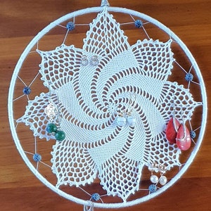 以下が含まれることがあります： 青いビーズと青いクリスタルがトップからぶら下がっている白いクロシェのドリームキャッチャー。ドリームキャッチャーは、緑、白、赤のイヤリングなど、さまざまなイヤリングで飾られています。