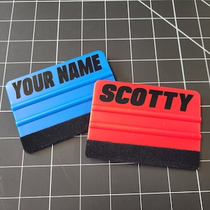 Peut inclure: Deux raclettes en plastique bleu et rouge avec des bords en feutre noir. La raclette bleue porte l'inscription "YOUR NAME". La raclette rouge porte l'inscription "SCOTTY".