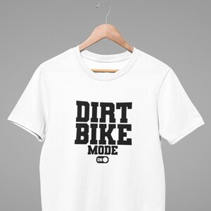 Może przedstawiać: Biała koszulka z czarnym napisem "Dirt Bike Mode ON" z grafiką przełącznika.