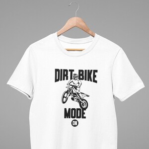 Może przedstawiać: Biała koszulka z czarnym nadrukiem przedstawiającym osobę jadącą na motocyklu terenowym. Pod nadrukiem znajduje się napis "Dirt Bike Mode On".