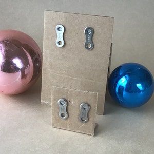 May include: Two pairs of silver earrings shaped like bike chain links displayed on a brown cardboard background. The earrings are displayed on two separate cards.