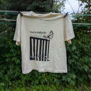 May include: A cream-coloured t-shirt featuring the phrase "Viva la revolución" and a graphic of a butterfly breaking free from a barbed wire fence. The t-shirt is hanging on a washing line with pegs.