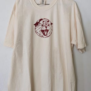May include: A cream colored t-shirt with a red graphic of a cat wearing a bandana with fish bones printed on it. The cat is looking up and has its mouth open. The text "COMFORT COLORS" is printed on the tag.