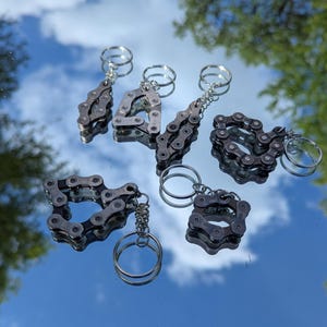 May include: A collection of six keychains made from repurposed bicycle chains. Each keychain features a unique shape, including teardrop, heart, and square designs. The keychains are silver-toned and have a reflective surface, with a silver key ring attached.