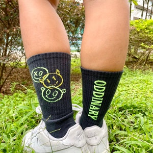 Może przedstawiać: Para czarnych skarpet z zielonym wzorem graficznym. Jedna skarpeta ma uśmiechniętą buźkę z napisem "ODDinary", a druga uśmiechniętą buźkę z napisem "XO". Skarpety są noszone z białymi butami sportowymi.