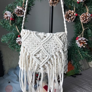 May include: White crocheted crossbody bag with a fringe detail. The bag is made of macrame and has a geometric pattern.