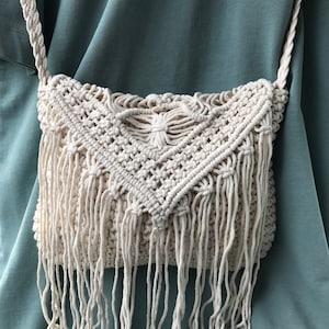 May include: Cream macrame crossbody bag with a woven pattern and long fringe. The bag has a braided strap and a rectangular shape. The background is a solid teal colour.
