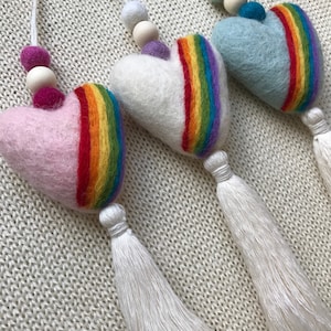 Może przedstawiać: Trzy filcowe ozdoby w kształcie serca z tęczowymi paskami i białymi frędzlami. Każde serce ma inny kolor: różowy, biały i jasnoniebieski. Serca są nawleczone na drewniane koraliki i małe filcowe kulki.