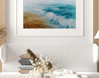 May include: A framed watercolor painting of ocean waves in shades of blue and white, crashing onto a sandy shore. The artwork hangs above a white sofa with decorative pillows and a bouquet of dried flowers.