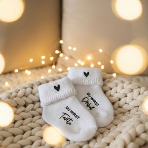 Könnte beinhalten: Ein Paar weiße Babysocken mit schwarzen Herzakzenten und deutschem Text. Die Socken liegen auf einer cremefarbenen Strickdecke, mit einem verschwommenen Hintergrund aus Kissen und Lichterketten.