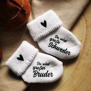 May include: White baby socks with black heart accents and the phrases "You will be a big sister" and "You will be a big brother" in black script. A brown baby shoe is visible in the background.