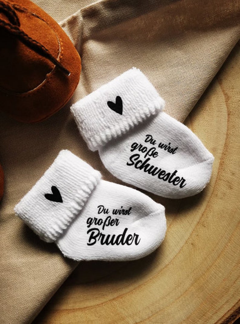 May include: White baby socks with black heart accents and the phrases "You will be a big sister" and "You will be a big brother" in black script. A brown baby shoe is visible in the background.