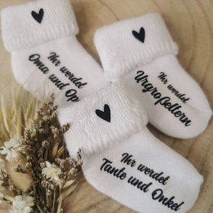May include: Three white baby socks with black text and a black heart. The socks are folded over at the top and have the phrases "Ihr werdet Oma und Opa", "Ihr werdet Urgro&szlig;eltern", and "Ihr werdet Tante und Onkel".