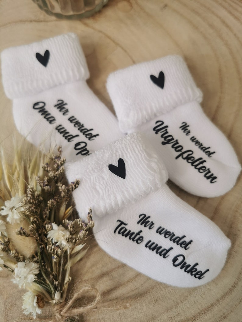 May include: Three white baby socks with black text and a black heart. The socks are folded over at the top and have the phrases "Ihr werdet Oma und Opa", "Ihr werdet Urgro&szlig;eltern", and "Ihr werdet Tante und Onkel".