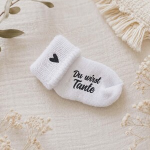 May include: White baby sock with a black heart and the text "Du wirst Tante". The sock is folded over, revealing a soft, textured fabric. The design is simple and elegant, perfect for a baby shower gift.