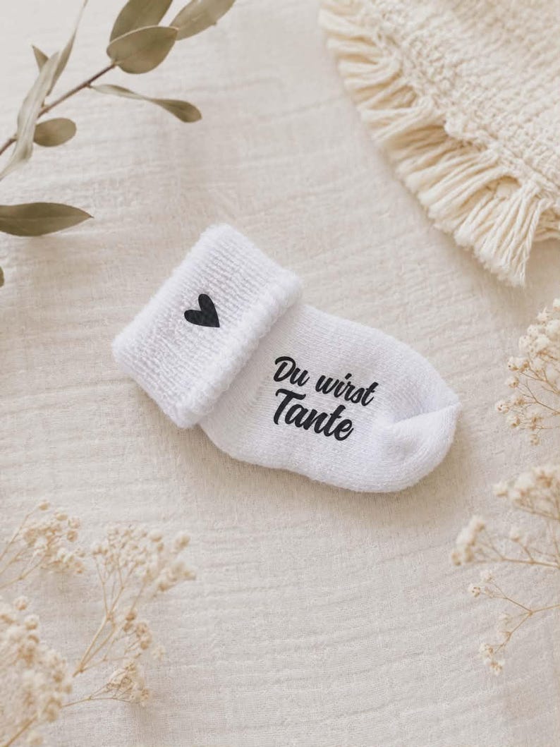 May include: White baby sock with a black heart and the text "Du wirst Tante". The sock is folded over, revealing a soft, textured fabric. The design is simple and elegant, perfect for a baby shower gift.