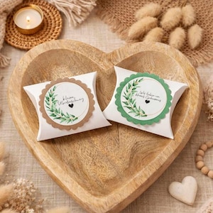 May include: Two white pillow boxes with scalloped edges, each featuring a circular label with botanical illustrations and text. The boxes rest in a heart-shaped wooden bowl, surrounded by decorative elements like a candle and a knitted blanket.