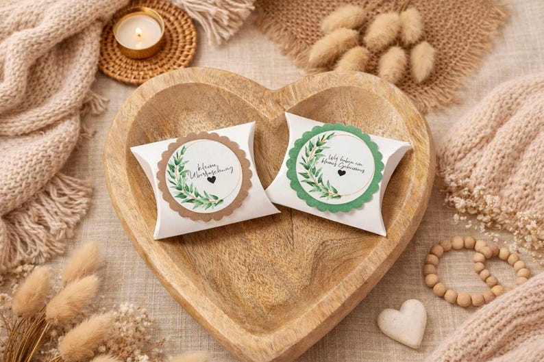 May include: Two white pillow boxes with scalloped edges, each featuring a circular label with botanical illustrations and text. The boxes rest in a heart-shaped wooden bowl, surrounded by decorative elements like a candle and a knitted blanket.