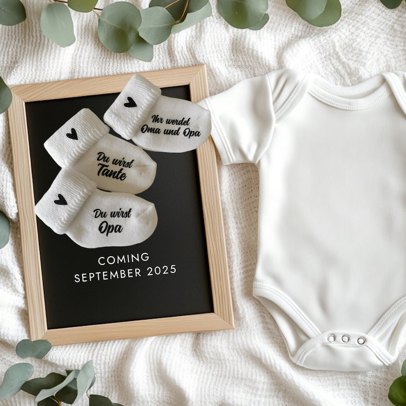 May include: A baby announcement featuring a framed chalkboard with white text and baby socks. The socks have black hearts and text that reads "Du wirst Tante", "Du wirst Opa", and "Ihr werdet Oma und Opa". The board says "COMING SEPTEMBER 2025". A white onesie is next to the board.