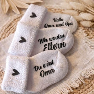 May include: White baby socks with black heart accents and text that reads "Hallo Oma und Opa", "Wir werden Eltern", and "Du wirst Oma". The socks are arranged on a neutral-toned surface with decorative elements.