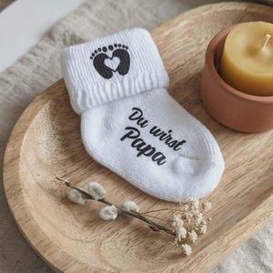 May include: White baby sock with the text "Du wirst Papa" and a footprint design with a heart. The sock is on a wooden tray with a candle and decorative sprigs.