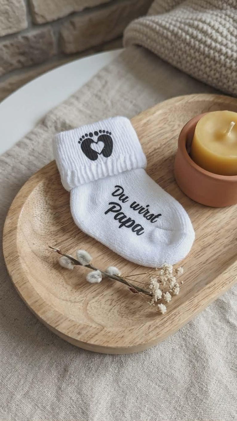 May include: White baby sock with the text "Du wirst Papa" and a footprint design with a heart. The sock is on a wooden tray with a candle and decorative sprigs.