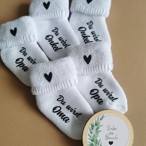 May include: White baby socks with black text that reads "Du wirst Opa", "Du wirst Oma", and "Du wirst Onkel", each with a black heart. A wooden disc with the text "Liebe im Bauch" and a green leaf design is also present.