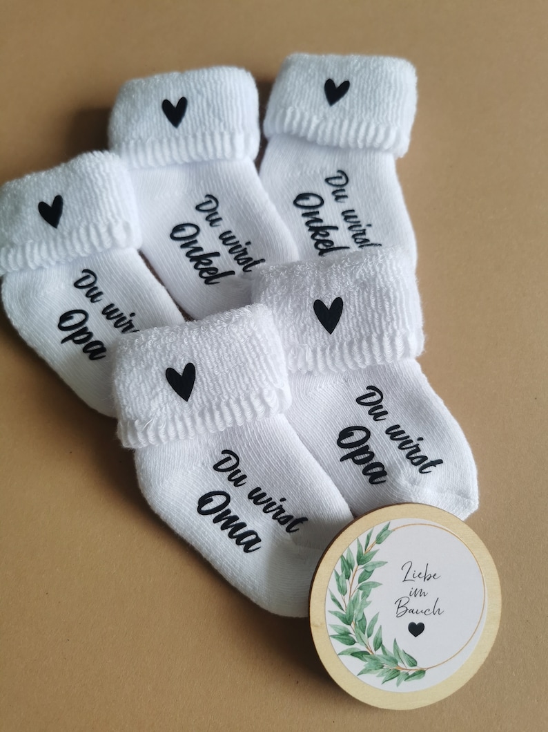 May include: White baby socks with black text that reads "Du wirst Opa", "Du wirst Oma", and "Du wirst Onkel", each with a black heart. A wooden disc with the text "Liebe im Bauch" and a green leaf design is also present.