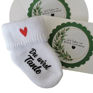 May include: White baby sock with a red heart and the words "Du wirst Tante" in black script. The sock is paired with a white gift box with a green scalloped edge and a circular design with text and greenery.