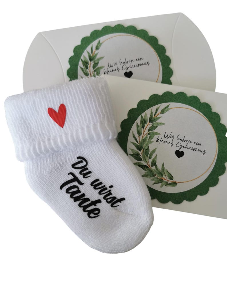 May include: White baby sock with a red heart and the words "Du wirst Tante" in black script. The sock is paired with a white gift box with a green scalloped edge and a circular design with text and greenery.