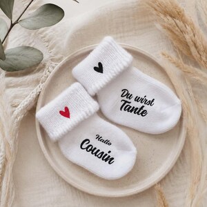 May include: Two white baby socks with rolled cuffs, each with a heart and text. One sock has a red heart and the words "Hallo Cousin", the other has a black heart and the words "Du wirst Tante". The socks are on a beige plate.