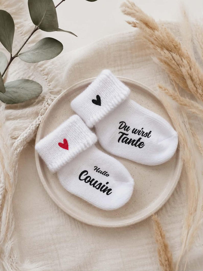 May include: Two white baby socks with rolled cuffs, each with a heart and text. One sock has a red heart and the words "Hallo Cousin", the other has a black heart and the words "Du wirst Tante". The socks are on a beige plate.