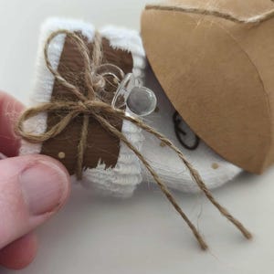 May include: A small, wrapped gift with a brown and white design, tied with twine. A clear dummy rests on top. The gift is next to a brown paper box, also tied with twine. The overall theme is baby or infant.