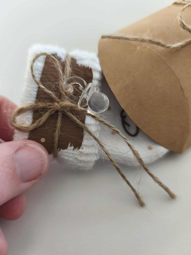May include: A small, wrapped gift with a brown and white design, tied with twine. A clear dummy rests on top. The gift is next to a brown paper box, also tied with twine. The overall theme is baby or infant.
