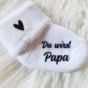 May include: White baby sock with a black heart and the words "Du wirst Papa" printed in black. The sock has a folded cuff and is made of a soft material. A baby item.