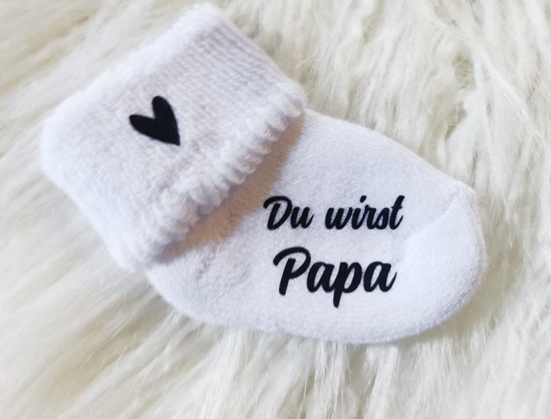 May include: White baby sock with a black heart and the words "Du wirst Papa" printed in black. The sock has a folded cuff and is made of a soft material. A baby item.