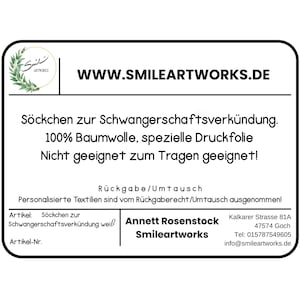 May include: A white rectangular label with rounded corners, featuring the text "WWW.SMILEARTWORKS.DE" and other German text. The label includes the Smileartworks logo and contact information. The text describes the product as socks for announcing pregnancy.