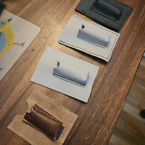 May include: A collection of leather cardholders in various colors, including gray, dark green, and brown. Each cardholder features a rectangular design with a curved cutout and a stitched accent. The cardholders are displayed on a wooden surface.