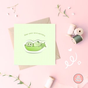 May include: A green card with a light green background and a white grid pattern. The card has the text "Hap-pea anniversary" in black text. There is a sticker of two green peas in a pea pod with faces and arms. The sticker is on a white background.