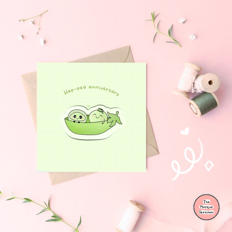 May include: A green card with a light green background and a white grid pattern. The card has the text "Hap-pea anniversary" in black text. There is a sticker of two green peas in a pea pod with faces and arms. The sticker is on a white background.